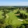 Parcours de golf verdoyant vue du dessus, avec fairways sinueux, plans d’eau et joueurs sur le green, entouré de arbres et un clubhouse au loin. Organiser un tournoi golf corporate.