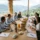 Une table ronde au sommet d’une serre lumineuse avec vue sur les montagnes, autour de laquelle des professionnels prennent le petit-déjeuner ou le brunch, documents et vaisselle visibles sur la table. Séminaire entreprise tout compris.