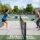 Deux femmes jouent au pickleball sur un court extérieur, se faisant face près du filet sur une ligne blanche; l’une frappe la balle avec une raquette bleue, l’autre réagit avec une raquette verte.