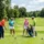 Quatre golfeurs sur un parcours verdoyant discutent du coup, le joueur en orange prête son driver tandis que les autres observent près du chariot. Formule de jeu golf shamble.