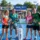 Quatre joueurs de padel souriants posent sur un terrain bleu lors d’un tournoi padel loisir, avec structures et public en arrière-plan.