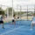 Quatre joueurs sur un court de padel en plein air, échange de balles près du filet. Le cadre est clôturé, ambiance sportive et conviviale. Le padel est-il un sport de riches?