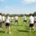 Groupe d’employés en tee-shirts blancs et shorts bleus jouant à un sport extérieur sur pelouse, lançant ou attrapant un ballon rouge lors d’un séminaire d’entreprise. Idées animations sportives séminaire entreprise.