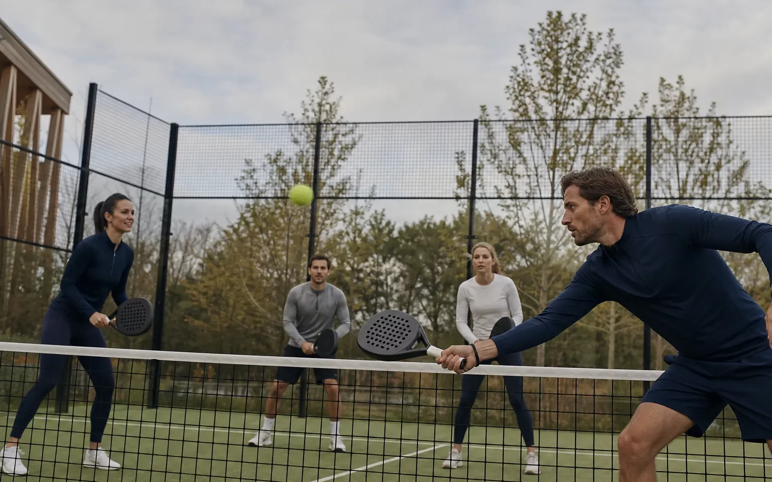 Quatre personnes jouent au tennis sur un court extérieur, deux près du filet et deux à l’arrière, en mouvement et concentrées.