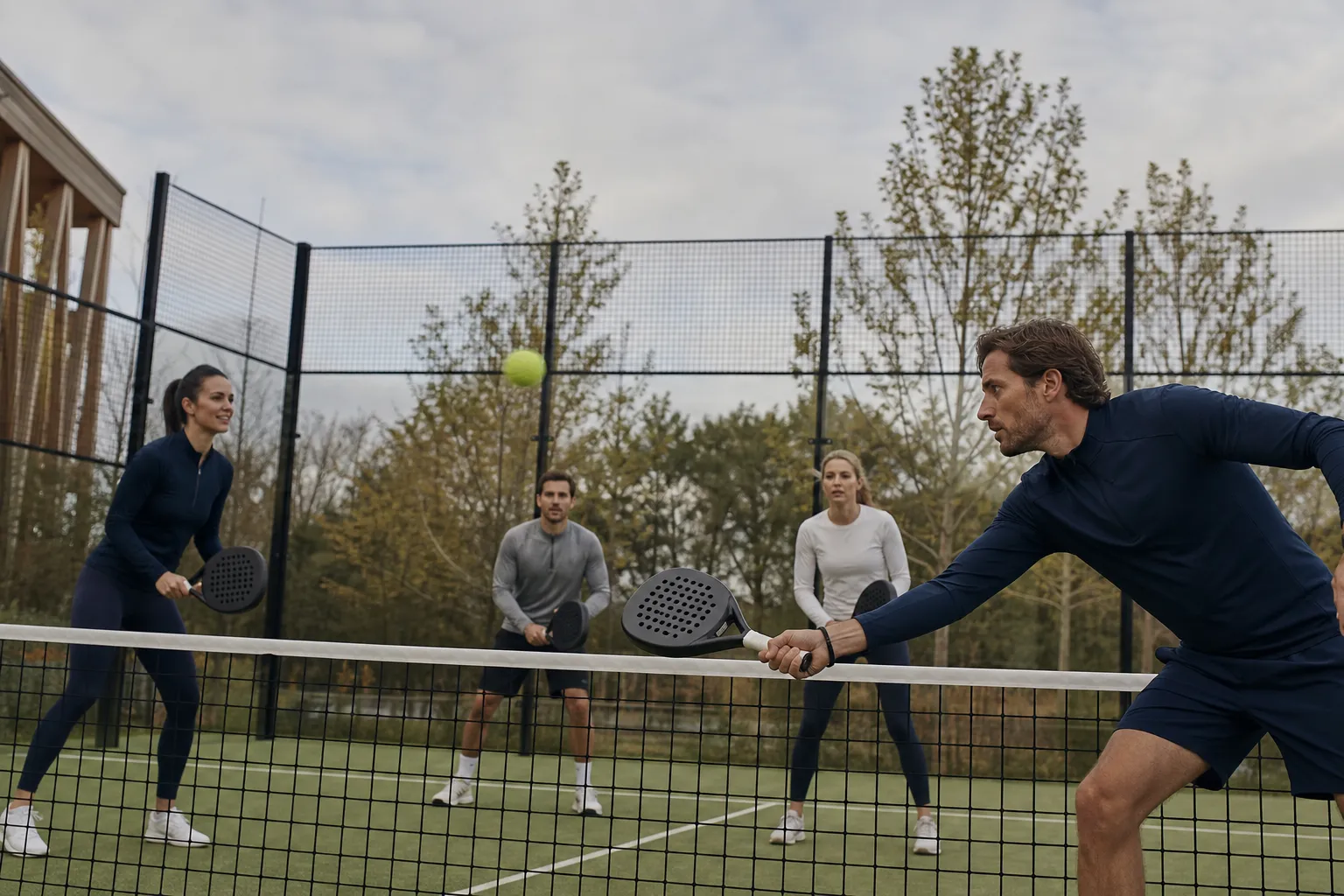 Quatre personnes jouent au tennis sur un court extérieur, deux près du filet et deux à l’arrière, en mouvement et concentrées.