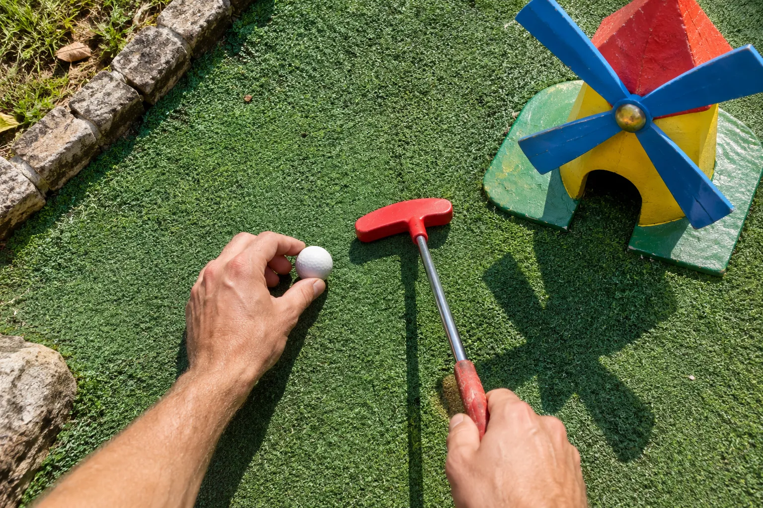 Personne tenant une balle blanche et utilisant un maillet rouge près d’un petit moulin en bois coloré sur une surface verte